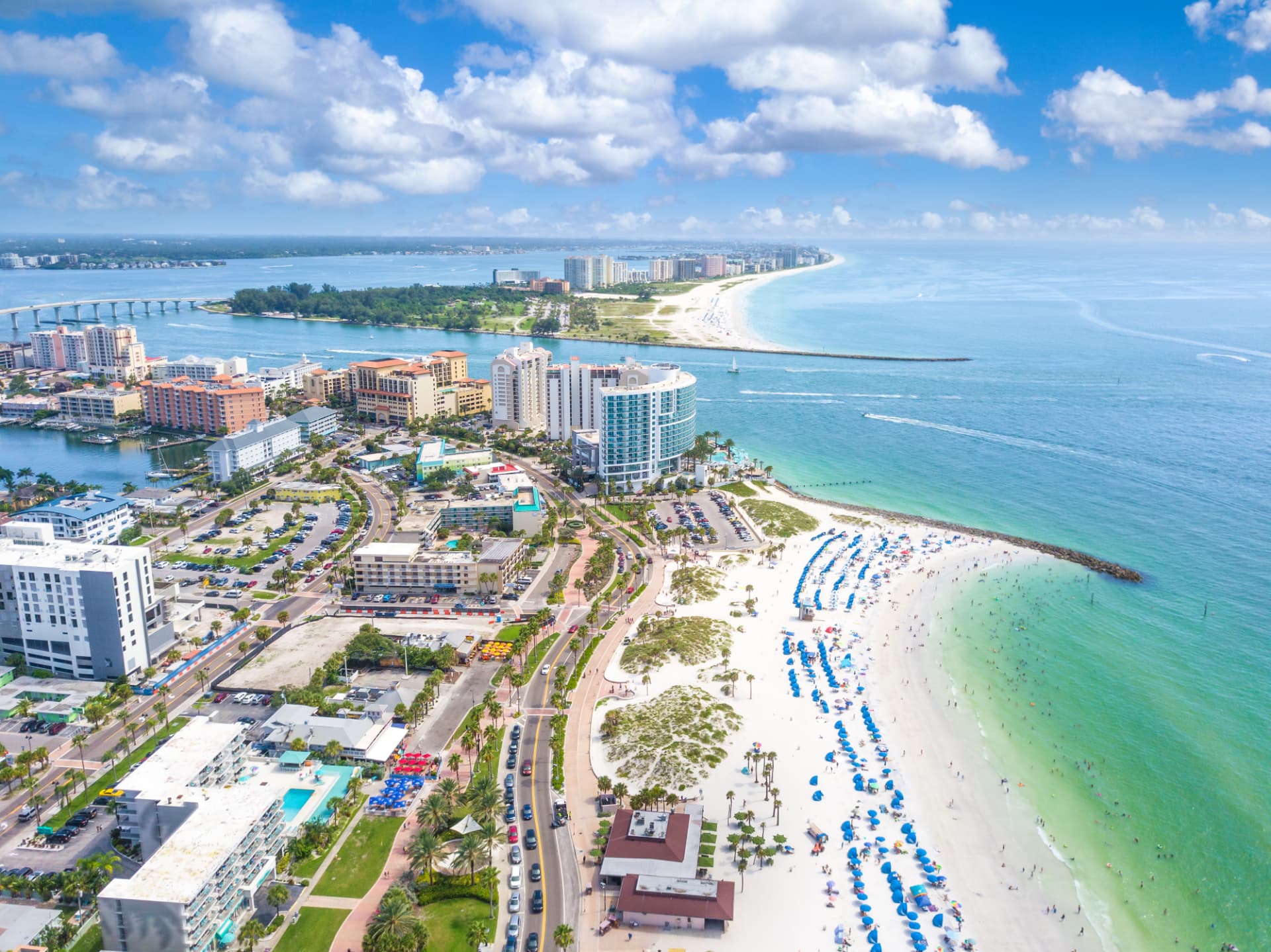 Daytime aerial view of Clearwater Beach, Florida, showing waterfront condominium and hotel buildings, a coastal roadway, white sand and the Gulf shoreline